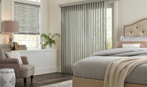 Modern bedroom with white vertical window blinds, gray bedding, and gray chair.