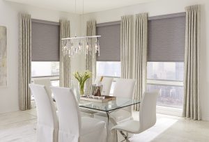 Luxurious dining area with plush seating, glass dining table, and three windows featuring roller shades.