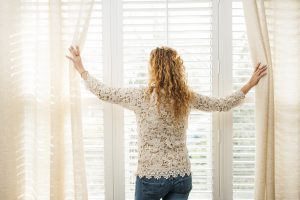 Picture of woman holding back a set of window drapes to look through her horizontal blinds.